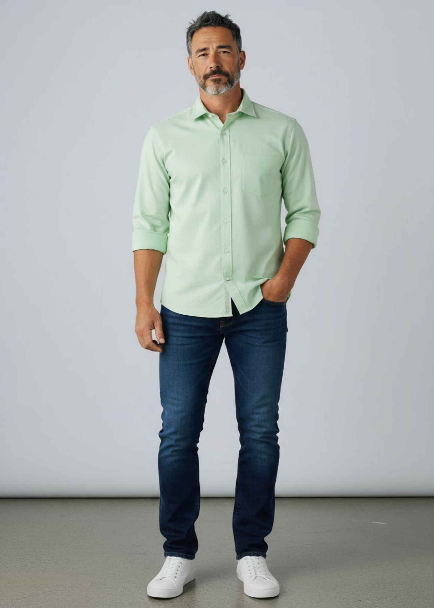 Man wearing a light green shirt, dark blue jeans, and white sneakers against a plain background