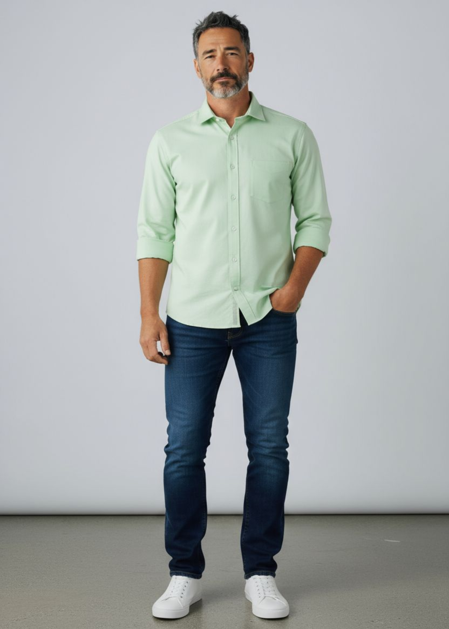 Man wearing a light green shirt, dark blue jeans, and white sneakers against a plain background